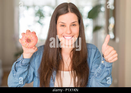 Hungrige junge Frau mit rosa Donut mit Lächeln tun ok Zeichen glücklich, Daumen hoch, mit den Fingern, ausgezeichnete Zeichen Stockfoto