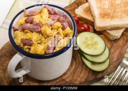 Rührei mit gebratenem Speck in einem Emaille Tasse, serviert mit getoasteten Scheibe Brot, Gurkenscheiben und Pflaume Tomaten auf Holzbrett Stockfoto