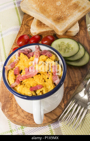 Rührei mit gebratenem Speck in einem Emaille Tasse, serviert mit getoasteten Scheibe Brot, Gurkenscheiben und Pflaume Tomaten auf Holzbrett Stockfoto