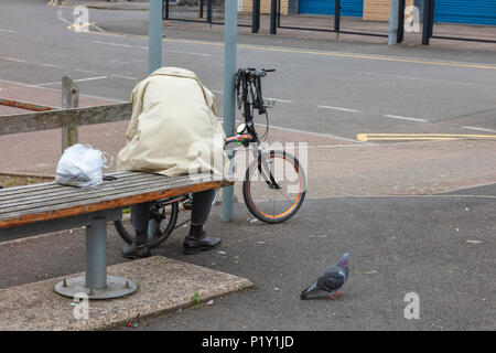 Eine ältere männliche Radfahrer sitzt auf einer Bank in Penarth, und versucht, eine defekte Bremse Kabel zu reparieren. Eine Taube Uhren auf. Cardiff, Wales, Großbritannien Stockfoto