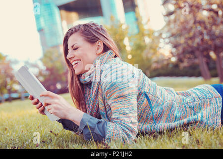 Junge Frau mit Tablet-PC im freien Festlegung auf Gras in einem Stadtpark, lächelnd. Stockfoto