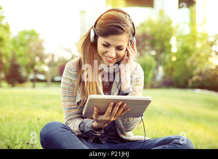 Lächelnde Frau Hören von Musik mit Tablet und Kopfhörer sitzen im Freien in einem Stadtpark Stockfoto