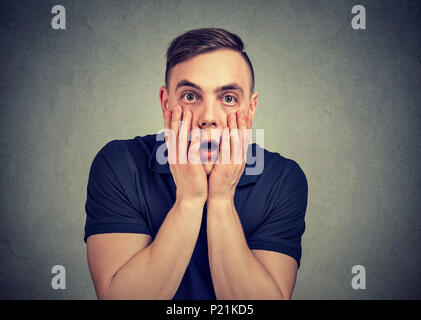 Junge legerer Mann berühren Gesicht in Verzweiflung posiert auf grau und Kamera mit Angst. Stockfoto