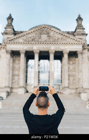 Nahaufnahme eines jungen kaukasischen Mann, von hinten gesehen, ein Bild von der Fassade des Reichstagsgebäudes in Berlin, Deutschland, mit seinem Smartphone Stockfoto