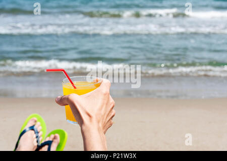 Nahaufnahme eines jungen kaukasischen Mann entspannt am Strand, tragen ein Paar der Grünen flip-flops, mit einem Glas mit einem erfrischenden orange Drink in seine Hände n Stockfoto