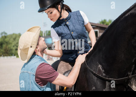 Attraktive Frau in Jockey Helm sitzen auf dem Pferd und Suchen auf den jungen Mann in Cowboy Hut Stockfoto