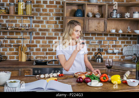Schöne junge Frau trinkt Wein beim Kochen Pizza in der Küche Stockfoto
