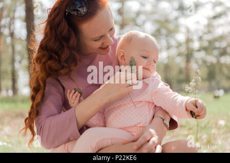 Das Mädchen, das kitzelt Wange des Baby, sitzen im Park, glücklich Lachen. Meine Tochter hält zwei Kegel in die Hände und schaut auf den Boden Stockfoto