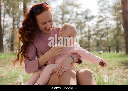 Das Mädchen, das kitzelt Wange des Baby, sitzen im Park, glücklich Lachen. Meine Tochter hält zwei Kegel in die Hände und schaut auf den Boden Stockfoto