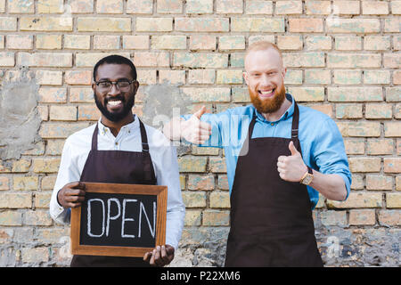 Stattliche multiethnischen Besitzer der Coffee Shop in Schürzen holding Zeichen geöffnet und zeigt den Daumen nach oben, in die Kamera lächeln Stockfoto