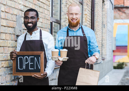 Junge schöne multiethnischen Besitzer der Coffee Shop in Schürzen mit Zeichen öffnen, Papiertüten und Einweg Kaffeetassen Lächeln an Kamera Stockfoto