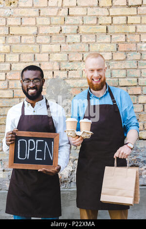 Jungen multiethnischen Baristas mit Vorzeichen öffnen, Papiertüten und Einweg Kaffeetassen Lächeln an Kamera Stockfoto