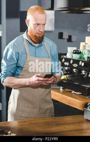 Bärtige junge Barista in Schürze mit Smartphone und Wegsehen im Coffee Shop Stockfoto