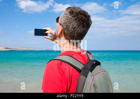 Ein Tourist am Strand nimmt ein Foto auf das türkisfarbene Wasser und die Küste mit seinem Handy Stockfoto