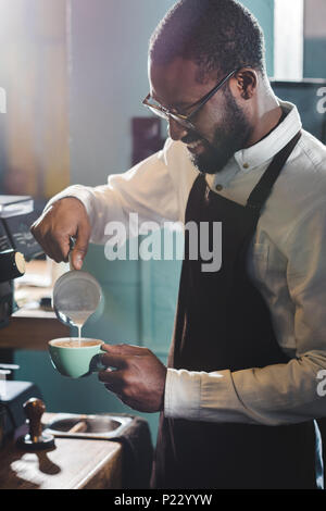 Lächelnden jungen afrikanischen amerikanischen Barista in Brillen gießen Milch, während sie Cappuccino Stockfoto