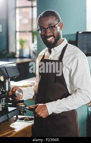 Junge afrikanische amerikanische Barista in Brillen Lächeln auf die Kamera während der Kaffee in der Kaffeemaschine Stockfoto