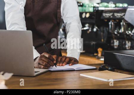 7/8 Schuß von jungen afrikanischen amerikanischen barista Notizen und mit Laptop im Coffee Shop Stockfoto
