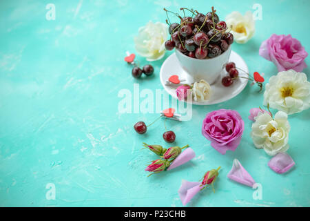 Weiße Tasse sweet cherry Beeren mit Rosen und Blumen auf grünem Smaragd Hintergrund. reifen Kirschen und wilde Rosen. Flach, Ansicht von oben süße Speisen floral background. Platz kopieren Stockfoto