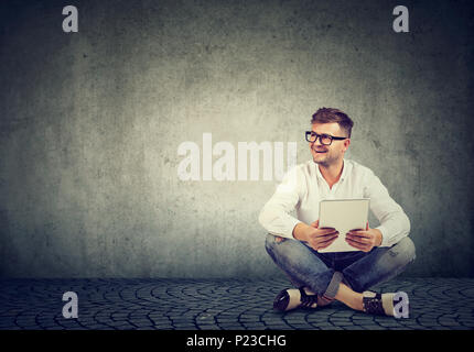 Fröhliche hipster Mann in Brillen und stylisches Outfit sitzen auf dem Boden gegen grungy Wand und mit Tablet. Stockfoto