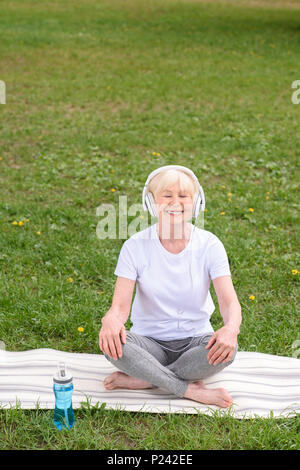 Gerne ältere Frau Musik hören mit Kopfhörern beim Sitzen auf Yoga Matte in Park Stockfoto