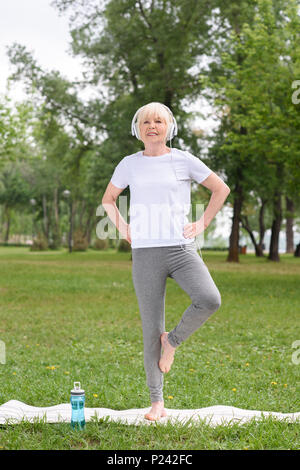 Ältere weibliche Yogi in Kopfhörern Üben Yoga Matte in Park Stockfoto