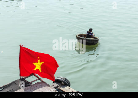 Phan Thiet, Vietnam - Januar 2014: Der Mann, der in den traditionellen Warenkorb Boot und vietnamesische Fahne auf dem Fluss in Phan Thiet, Vietnam Stockfoto