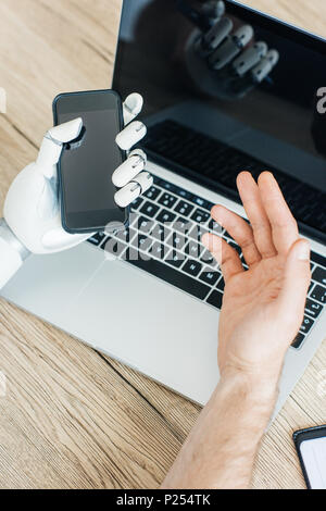 Nahaufnahme von Mensch und Roboter Hand mit Smartphone und Laptop an den hölzernen Tisch Stockfoto