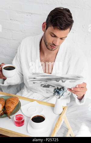 Jungen gutaussehenden Mann im Bademantel lesen Zeitung beim Frühstück mit Kaffee und Croissants. Stockfoto