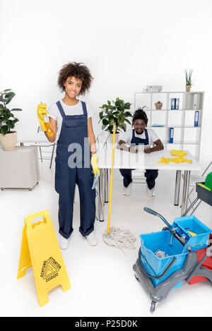 Schöne junge Reiniger, Sprühflasche Lächeln auf die Kamera während der männlichen Kollegen mit digitalen Tablet hinter Stockfoto