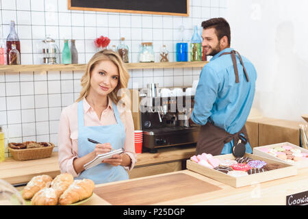 Schöne junge Kellnerin Notizen und lächelnd an der Kamera, während Barista hinter dem Bilden Stockfoto