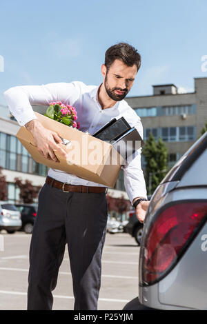 Junge Manager mit Box von persönlichen Sachen öffnung Kofferraum auf Parkplatz Stockfoto