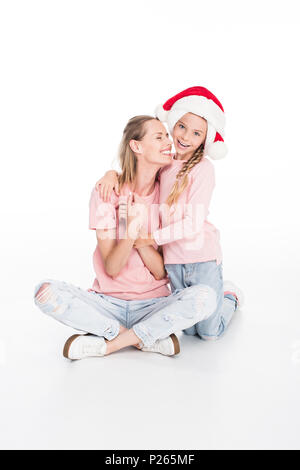 Lächelnde Frau und Tochter tragen Nikolausmützen sitzen auf dem Boden umarmen, Stockfoto