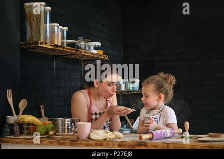 Junge Mutter und Tochter bereiten Sie Cookies in der Küche. Sie sind in den Schürzen. Frau blasen Mehl in Ihre Hände. Mädchen sind Aprilscherze. Zeit mit der Familie. Stockfoto