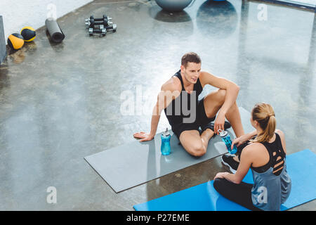 Hohe Betrachtungswinkel der Sportler und Sportlerin mit Flaschen Wasser sitzen auf Yogamatten und an jedem anderen Suchen in der Turnhalle Stockfoto