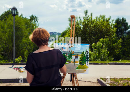 Eine blonde junge Künstlerin in einem schwarzen T-Shirt und Jeans Farben auf Leinwand und einer Staffelei mit Ölfarben in einem Stadtpark an einem sonnigen Tag. Zeichnung auf einer o Stockfoto