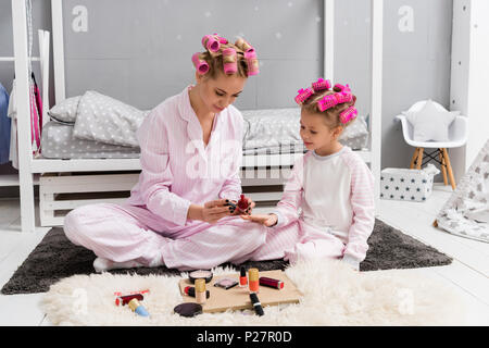 Junge Mutter Malerei Nägel der Tochter mit polnischen, während Sie sitzen im Schlafanzug mit Haar Rollen Stockfoto