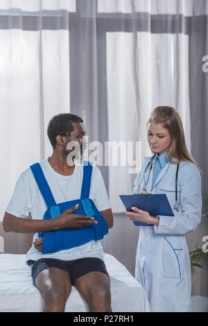 Junger Arzt schriftlich etwas in die Zwischenablage über Afrikanische amerikanische Patienten mit Arm Brace Stockfoto