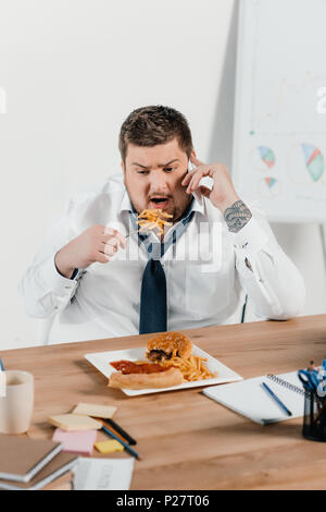 Übergewicht Geschäftsmann sprechen auf dem Smartphone während Junk food am Arbeitsplatz Essen Stockfoto