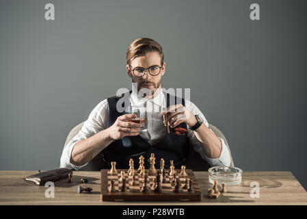 Junge Unternehmer mit einem Glas Whisky und Zigarre auf Kamera beim Schach spielen Stockfoto