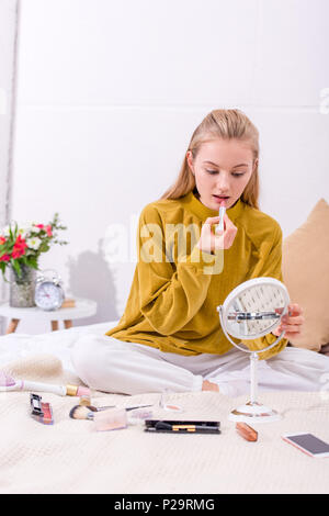Junge Frau Anwendung Lippenstift auf Bett zu Hause und Spiegel Stockfoto
