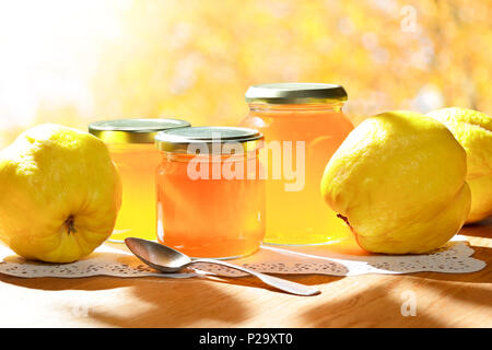 Quittengelee in Gläsern mit Quitten auf einem Holztisch in strahlendem Sonnenschein vor einem gelben und orangenen Hintergrund. Stockfoto