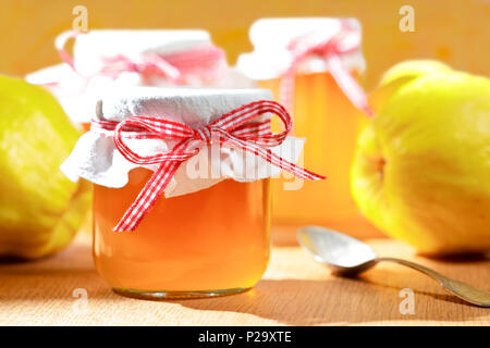 Quittengelee in Gläsern mit Quitten auf einem Holztisch in strahlendem Sonnenschein vor einem gelben und orangenen Hintergrund. Stockfoto