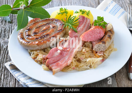 Bauch von Schweinefleisch und verschiedenen Würstchen auf Sauerkraut mit gekochten Kartoffeln, alle auf einem alten Holztisch Stockfoto