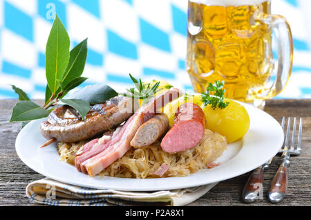 Bauch von Schweinefleisch und verschiedenen Würstchen auf Sauerkraut mit gekochten Kartoffeln, alle auf einem alten Holztisch, im Hintergrund eine Bayerische Fahne Stockfoto
