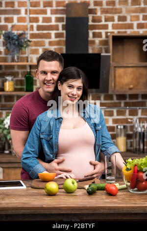 Glückliche junge schwangere Paar umarmen und lächelnd an der Kamera, während zusammen kochen Stockfoto