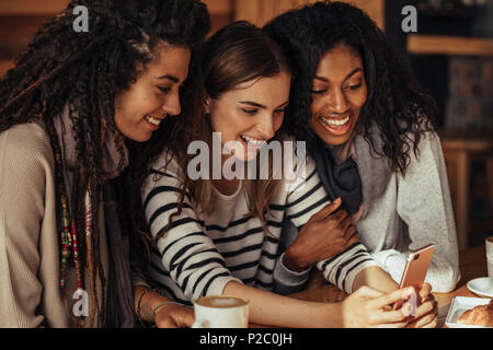 Drei Frauen in einem Restaurant mit Blick auf Handy sitzen und Lachen. Freunde sitzen in einem Café mit Kaffee und Snacks auf den Tisch mit einem m Stockfoto