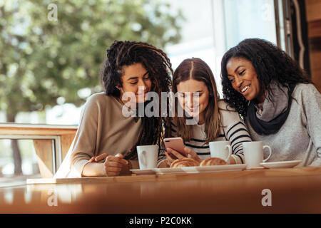 Drei Frauen sitzen in einem Restaurant am Handy suchen und lächelnd. Freunde in einem Café mit Kaffee und Snacks auf den Tisch in einem mo Suchen sitzen Stockfoto