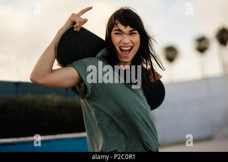 Portrait von aufgeregt Frau Skateboarder im Freien an Skate Park. Frau mit ihrem Skate Board bei Kamera schaut und lacht im Freien. Stockfoto