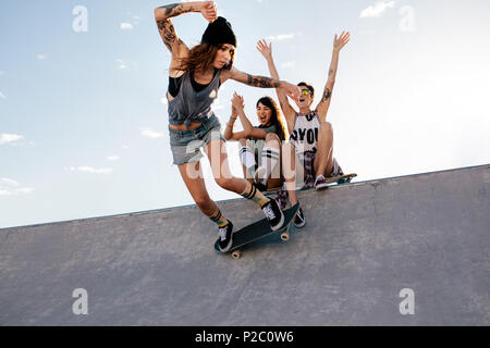 Skater girl reitet auf Skateboard mit weiblichen Freunde sitzen auf Rampe zujubeln. Weibliche skateboarding bei Skate Park mit Freunden zujubeln. Stockfoto