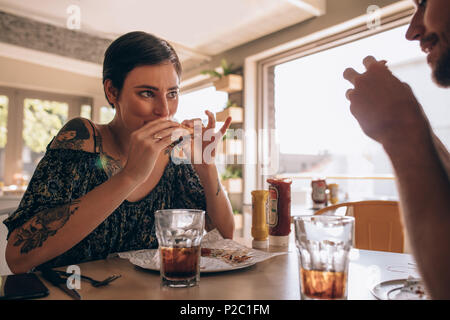 Junge Frau essen Burger mit ihrem Freund sitzen um einen Tisch in einem Restaurant. Paar Burger in das Cafe. Stockfoto